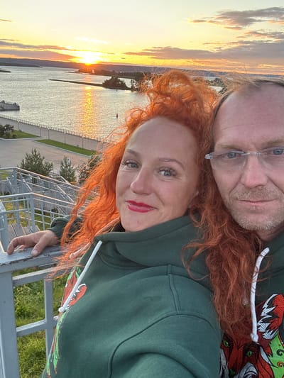 The image is a selfie taken by two individuals, a man and a woman, with bright red hair. They are wearing matching dark green hooded sweatshirts with a colorful graphic of a dragon. The man, on the top of the frame, wears glasses and has stubble. The woman, below him, has red lipstick and blue eyes. They are positioned in close proximity, with the woman's head tilted upwards and the man's head tilted downwards.

The background shows a body of water, likely a river, with the sun setting or rising, casting a golden glow on the clouds and water. The sky is filled with dramatic clouds, illuminated by the warm hues of twilight. In the distance, a line of trees and a shoreline are visible, along with some structures that appear to be part of a walkway or observation deck. The overall mood seems to be romantic and serene, with the couple enjoying the scenic sunset/sunrise. The image is oriented vertically, as if held up for a selfie. No visible text is present in the image. The setting appears to be outdoors, possibly near a waterfront.