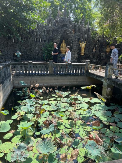In Cam Ranh, Vietnam, at a location that appears to be a Buddhist temple, a pond filled with lotus pads and wilting flowers is visible. A stone wall with numerous small statues and a few larger golden Buddha statues is behind the pond. A concrete bridge with a railing spans the pond. Three individuals are present: a man in a black t-shirt and shorts stands on the left side of the bridge, holding a bottle of water; a woman in a white top and shorts walks across the bridge; and a young man in a patterned shirt and pants walks away from the camera on the right side of the bridge. The scene is bathed in bright sunlight, casting shadows on the water and lotus pads.