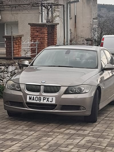 A light brown/champagne-colored BMW E90 3 Series sedan, registered with the British license plate MA08 PXJ, is parked outdoors on a paved surface in the city of Topolovgrad, Bulgaria. The pavement consists of light-colored, rectangular pavers arranged in a herringbone pattern. The vehicle is positioned facing the viewer, with its front bumper, grille, headlights, and part of the hood and windshield clearly visible. The interior of the car is not clearly discernible through the tinted front windshield.
In the background, behind the BMW, is a weathered, two-story building with a light-colored, stucco or rendered facade. Two reddish-brown brick pillars with light-colored caps are visible on the left, framing a white metal gate or fence. Bare tree branches, possibly from a trained vine, extend across the top of the frame. To the right, a white panel van is partially visible behind the BMW's roofline. The distant background features a dark, possibly tree-covered hillside under an overcast sky. No individuals are present in the frame.