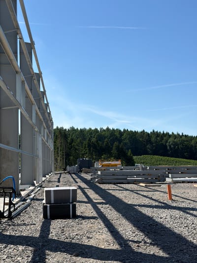 The image depicts an outdoor construction site with a large, unfinished building structure on the left. The building's framework consists of prominent gray metal beams and supports, forming a tall, linear wall. The ground is covered in gravel, with long, distinct shadows cast by the building's structure, indicating a sunny day. In the foreground, two dark rectangular objects are stacked, wrapped in white strapping. Further into the scene, there are piles of gray construction beams, a yellow piece of equipment resembling a lift or crane, and a black water tank. Beyond the construction area, a dense line of green trees forms a natural backdrop, with a bright blue sky above, marked by faint wispy clouds. The scene is consistent with an industrial or commercial construction project in a semi-rural or wooded area.