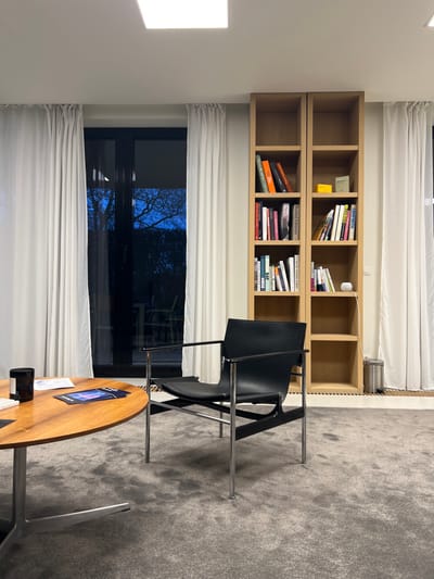 A room contains a bookshelf filled with books, a large window obscured by white curtains, and two black chairs arranged around a round wooden table. The room is carpeted with gray material. Outside the window, dark trees are visible. A small black mug sits on the table, next to a stack of white papers.
