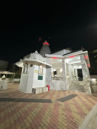 At night, an illuminated multi-story Hindu temple in Vadodara, India. The structure is predominantly white, with some red and green accents. The ground level is paved with red and yellow bricks in a patterned design. The background is a dark sky.