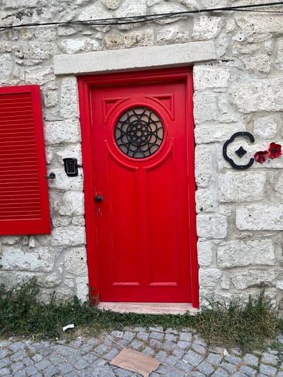 The image displays the exterior of a building in Çeşme, Turkey, featuring a vibrant red door and an adjacent, partially open red louvered shutter set within a light-colored, rough-hewn stone wall. The red door, centrally located, includes a circular window with an intricate black metal grilwork design and is equipped with a black doorknob and keyhole. Above the door, a light-colored stone lintel is visible. To the immediate left of the door, a black electronic keypad access control device is mounted on the stone wall. To the right of the door, a decorative black metal "C" shape, containing a smaller black diamond element, is affixed to the wall, with two artificial red poppy flowers attached to its right side. Black electrical wires run horizontally along the stone wall above the door and shutter. The ground in the foreground consists of uneven grey and tan cobblestone pavement, with green weeds and grass growing along the base of the wall and between some cobblestones. A small piece of white debris and a flat piece of cardboard are present on the cobblestones. No individuals are visible in the scene.