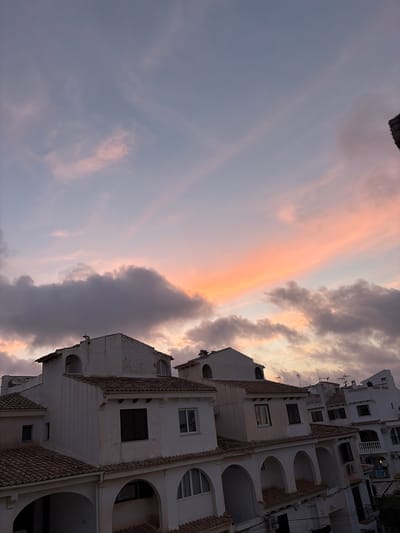 This media file depicts a building in Torrevieja, Spain, against a backdrop of a sunset sky. The building is white with multiple stories, featuring balconies with arched openings and tiled roofs. Various windows are visible, some with shutters. The sky displays a gradient of colors, with soft pinks and oranges from the setting sun blending into darker grays and blues of the clouds. The scene suggests a tranquil evening. No people are visible in the frame. No text is discernible. The image is oriented vertically.