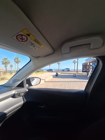 The image is taken from inside a car, looking out through the windshield and side window. The perspective suggests the car is parked or moving slowly. The sun is shining brightly, indicating it is daytime. The scene outside the car is a coastal area with palm trees and the sea in the background.
In the foreground, inside the car, is the sun visor with an "AIRBAG" warning label. The label features symbols indicating safety precautions for airbags, including a child in a car seat and a warning not to place objects or children in front of the airbag. The interior of the car is dimly lit in some areas and brightly lit in others, reflecting the outdoor sunlight.
Outside the car, a paved walkway lines the road, with a line of parked cars and a food truck visible. The food truck has signage that is partially obscured but appears to be for a business named "SANTIS". Beyond the walkway and cars, the blue sea stretches out, meeting a clear blue sky. The presence of palm trees and the visible signage suggest a location in a warm climate, consistent with Safi, Morocco. The overall impression is a bright, sunny day in a coastal town, viewed from the comfortable interior of a vehicle.
