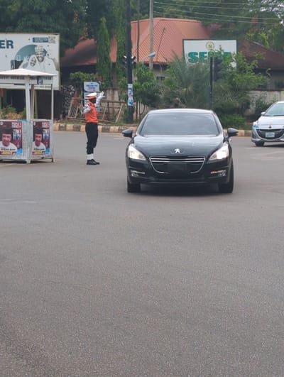 The image shows a black Peugeot car traveling down a street. A uniformed traffic officer is directing traffic. Political campaign posters are visible. A sign for "Thelish Eye Centre" can be seen in the background. There is also a sign for "SER." This appears to be in Shaba, Nigeria. Other cars are on the street. The environment includes trees and buildings.