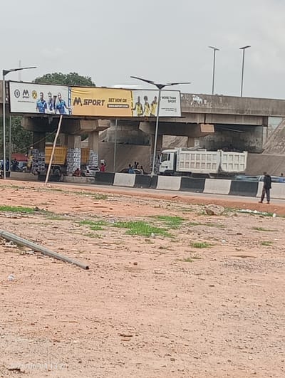 The image captures an outdoor scene beneath a concrete overpass, suggestive of a commercial or transit area, likely in Madalla, Nigeria. A road runs under the overpass, flanked by dirt and gravel foreground.

Two large billboards are affixed to the underside of the overpass. The left billboard displays the "M SPORT" logo alongside a "BVB SPORT" emblem, featuring male athletes in sports jerseys. Prominent text reads "BET NOW ON WWW.MSPORT.COM". The right billboard also shows an "M SPORT" logo and the slogan "MORE THAN SPORT," with more images of athletes.

On the left side of the road beneath the overpass, a yellow dump truck is parked, loaded with numerous packaged goods, which appear to be white jerry cans. Several people are visible around this truck, some sitting, others standing. A white dump truck is also seen on the right side of the road, driving away from the camera. Further right, a person in dark attire walks along the roadside.

The foreground consists of reddish-brown, uneven dirt and gravel, with sporadic patches of green vegetation. A metal pole or pipe lies horizontally in the lower left. The sky is uniformly light and overcast. A timestamp in the lower left corner indicates "2026/06/04 13:20," suggesting the photograph was taken in the early afternoon. The scene conveys a sense of bustling activity in a functional, semi-urban setting.