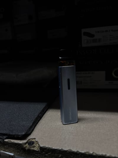 A silver vape device is placed on a gray surface. The device is rectangular with a golden cap on top. The word "VAPORESSO" is visible on the side of the device. The background is dark and out of focus, with some text visible on what appears to be packaging. The lighting suggests an indoor environment. No information about Zhytomyr, Ukraine is present in the image.