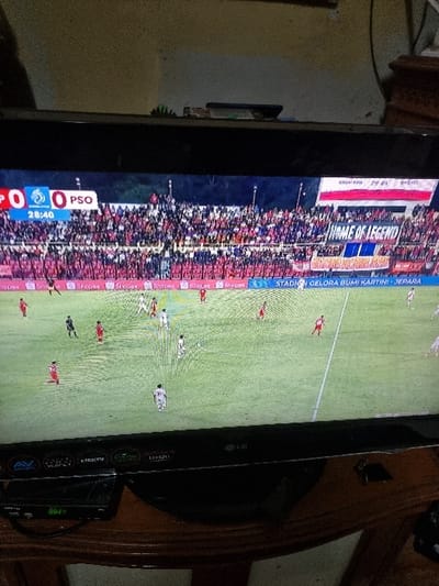 The image depicts an LG brand flat-screen television displaying a soccer match, situated within an indoor residential setting in Pacet, Indonesia.
The television screen shows a live soccer game on a green pitch. The scoreboard overlay at the top left indicates a score of "0 0 PSO" and a game time of "28:40". Players are visible on the field, with one team wearing red jerseys and the other in white and black striped jerseys. In the background, a stadium filled with spectators, largely in red, is visible. A prominent banner across the stands reads "HOME OF LEGEND", and a digital advertising board below it states "STADION GELORA BUMI KARTINI - JEPARA", identifying the match venue. An Indonesian flag (red and white) is displayed at the stadium's upper level.
Below the television, a black electronic set-top box or media player is visible on a wooden surface, displaying the numerical value "0024" in green. The television's lower bezel shows labels for various inputs, including "AV", "VOICE", "HDMI", and "LG" branding. The television rests on a dark wooden table or stand with a curved edge. The background wall behind the television is light-colored and unadorned.