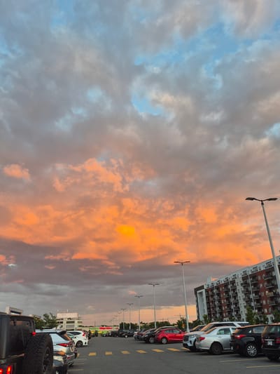 This is a photograph of a parking lot in Laval, Canada, taken at dusk. The sky is filled with dramatic clouds, painted with hues of orange, pink, and grey, with patches of blue sky visible. A modern apartment building with a red and white facade stands in the background on the right. Various cars are parked in the lot, including a Jeep in the foreground on the left. Several streetlights line the parking lot. The scene appears to be a commercial or residential area.