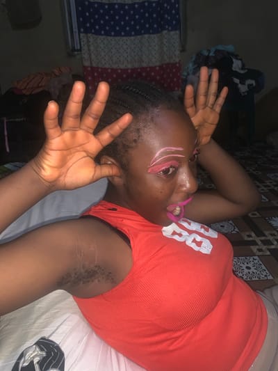 An African female, identified as being in Agban, Nigeria, is shown in a close-up, low-angle shot. She is wearing a vibrant orange tank top with a ribbed texture and a white graphic design across the chest. Her makeup is bold, featuring bright pink lipstick and eyeshadow, and stylized pink eyebrows. Her right hand is raised, fingers spread, near her face, while her left arm is bent, with her hand touching her ear. Visible on her right arm are several faint, light-colored stretch marks. The background is dark and somewhat blurred, with hints of a flag-like pattern on the wall, possibly a representation of the Nigerian flag, and what appears to be a pile of clothes or fabric. The floor has a patterned tile design. The overall impression is of a candid and expressive moment, likely captured indoors.