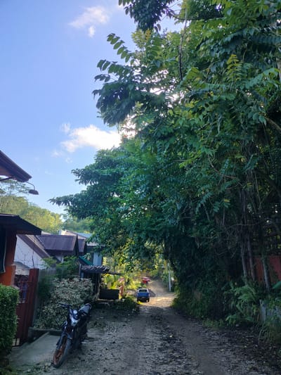 The image depicts a rural dirt road in Kidapawan City, Philippines, flanked by lush green foliage and residential structures. The sky is blue with scattered clouds, and the sun's rays are visible peeking through the leaves of a large tree on the right. On the left side of the road, several houses with corrugated metal roofs are visible. A motorcycle is parked in the foreground on the left. Further down the road, a blue pickup truck and two red vehicles are seen. The environment appears to be a residential area with a mix of natural vegetation and man-made structures.