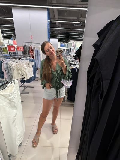 A young woman with long brown hair pulled into a ponytail takes a mirror selfie in a clothing store. She is wearing a green ruffled blouse and light blue denim shorts. She has one hand in her pocket and is holding a smartphone in the other. A disposable face mask with a floral pattern hangs from her other hand. Behind her, racks of clothing are visible, and the store's interior lighting illuminates the scene. The woman appears to be shopping in Novorossiysk, Russia, as indicated by the Cyrillic text on store signage. The floor is tiled in a light color. To the right of the frame, a black garment is hanging, partially obscuring the reflection.