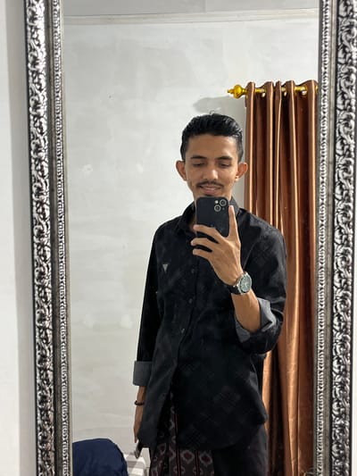 A male individual, appearing to be in an indoor setting in Alueduamuka O, Indonesia, is captured in a mirror selfie. He is observed from the waist up, facing the camera with a slight smile. He has dark, slicked-back hair and a thin mustache. He wears a black long-sleeved collared shirt with a subtle checkered or plaid pattern and a small, triangular logo on the left chest; the sleeves are rolled up at the cuffs. A silver-toned watch with a dark band is on his left wrist, and a dark beaded bracelet is on his right wrist. He holds a black smartphone in his right hand, positioned to capture the image. Below his shirt, a patterned, dark-colored sarong or traditional wrap is visible, with dark trousers underneath. The background visible in the mirror includes a plain white wall to the left and a brown curtain with a gold finial and rod to the right. The mirror itself has a wide, ornate, silver-colored frame. A dark blue object is partially visible on what appears to be a light-colored tiled floor in the lower left reflection.