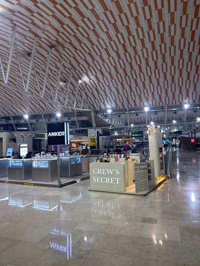 The image depicts the interior of a modern airport terminal, likely in a city with a large airport. The ceiling features a distinctive orange and white striped pattern supported by a metal framework. In the foreground, there is a retail display with the prominent sign "ANKER," suggesting a store selling electronics or related accessories. Adjacent to this, another display reads "CREW'S SECRET," possibly a duty-free shop or a store selling perfumes and cosmetics. Several glass display cases showcase various products. The floor is polished and reflective, with the lights from the displays creating bright reflections. In the background, airport signage and other shops are visible, along with a few blurry figures of people walking through the terminal. The overall impression is of a busy, well-lit commercial area within an airport. The location is not specified as Baji Mangngai, Indonesia; however, the presence of airport infrastructure suggests a travel hub.