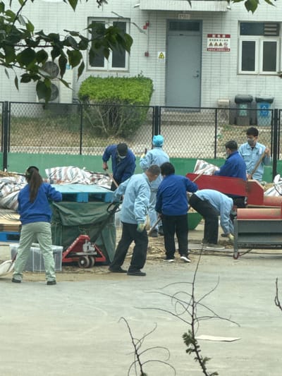 In Daxing District, China, a group of at least eight people are engaged in an outdoor task near a light-colored building with windows, an entrance door, and warning signs. The building also has air conditioning units installed on the wall. A chain-link fence separates the workers from the building. Some workers wear blue shirts, while others wear light blue shirts. Most have on dark pants, while at least one wears light-colored pants. They appear to be using tools near some equipment covered by a green tarp. A red cart is visible near the tarp. Branches and leaves are present in the foreground, partially obscuring the view.