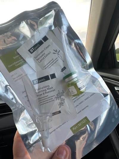 The image shows a sealed plastic packet containing skincare products. The packet is labeled "BTpeel" and includes individual packets of "Sunscreen Cream Recovery Carrot Extract", "Moisturizing Soothing Mask Rose & Beeswax", and a small green glass vial with a white cap. The packet is held in someone's hand, likely inside a car based on the interior visible in the background. The packet also shows writing in Russian. There is no information available in the image about Ryabinovka, Russia.