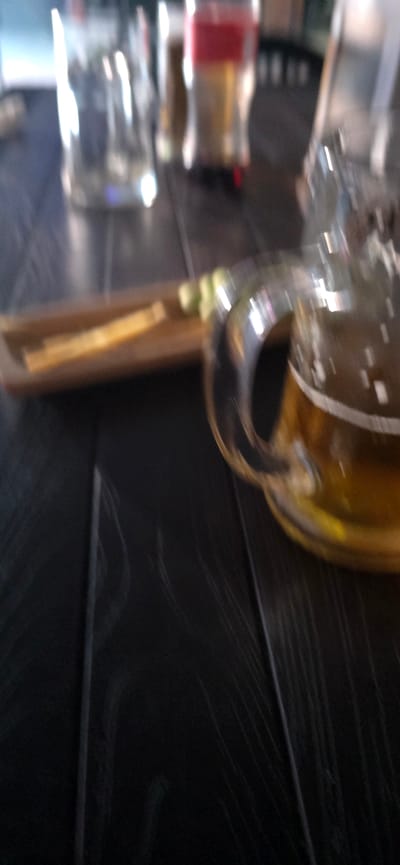 The image depicts a close-up, slightly blurred view of a table with drinks. In the foreground, a mug of beer with a frothy head is visible on the right. To the left of the beer mug, a wooden tray holds two pieces of what appear to be cheese and a garnish of lime or cucumber. In the background, out of focus, are several other glasses and bottles, some containing liquids of different colors. The table surface is dark and appears to be made of wood with visible grain. The lighting suggests an indoor setting, possibly a bar or restaurant. The specific location in the city of Gori, Georgia, is not discernible from the image itself.