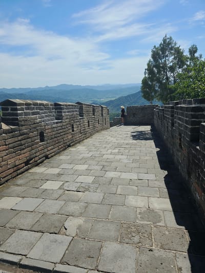 The image shows a section of the Great Wall of China, located in the Huairou District of China. The wall is constructed of dark bricks and paved with large stone slabs. A person wearing a wide-brimmed hat stands on the wall, looking out at the surrounding landscape of rolling hills and mountains. The sky is blue with scattered white clouds. The scene is set on a bright, sunny day.