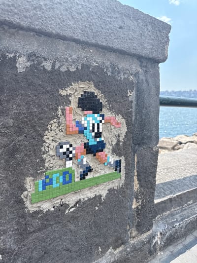 The image shows a pixelated mosaic of a cartoon character, possibly a video game character, adhered to a rough, dark gray concrete surface. The mosaic depicts a figure with dark hair, a blue and white checkered shirt, and brown and pink legs. The character appears to be in motion, with one arm extended and a green and blue checkered object beside it, possibly representing a ball or a background element. The mosaic is partially embedded in white grout or cement, which has been applied somewhat unevenly to the concrete.
The concrete surface appears to be part of a wall or ledge, with a textured, weathered appearance. Above and to the left of the mosaic, a bright blue sky with scattered white clouds is visible, suggesting it is daytime and likely fair weather. In the background, out of focus, is a body of water, possibly the sea, with light glinting on its surface. A hint of a railing or pipe can be seen above and to the left, near the sky. The overall scene suggests an outdoor urban or coastal environment, possibly in Naples, Italy, given the provided context. There is no visible text in the image. The mosaic artwork is the central focus, bringing a splash of color and whimsy to the otherwise utilitarian concrete structure.
