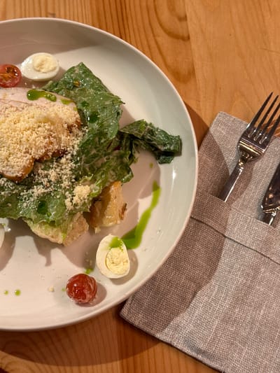 A close-up, high-angle shot captures a Caesar salad served on a white plate. The salad contains romaine lettuce, croutons, thinly sliced chicken, grated Parmesan cheese, a halved quail egg, and a cherry tomato. A drizzle of green dressing is visible on the plate and salad. The plate rests on a light-colored wooden surface. To the right of the plate, a gray linen napkin is folded, with a silver fork and knife tucked into it. The lighting casts a soft shadow on the napkin. There is no indication of the city of Bakhtiyary, Russia, in the image.