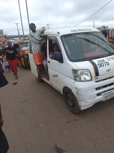 In Enugu, Nigeria, a white minibus is seen with its door open and a man standing on the threshold, his upper body outside the vehicle. He is wearing a light blue and white patterned long-sleeved shirt and orange shorts. Inside the bus, two passengers are visible. To the left of the minibus, a yellow and green auto-rickshaw is parked. Further to the left, several people are walking, including a woman in a black t-shirt and a red skirt, carrying a white bag. The scene appears to be a busy street or transportation hub, with overhead wires and a cloudy sky. The minibus has markings on its exterior, including "ENTRACO" on the side and "ENS/104/5770" on the top. The front of the minibus also displays "ABK R-109 9070" and a license plate reading "UWN-334XN". The minibus has visible damage to its front bumper.