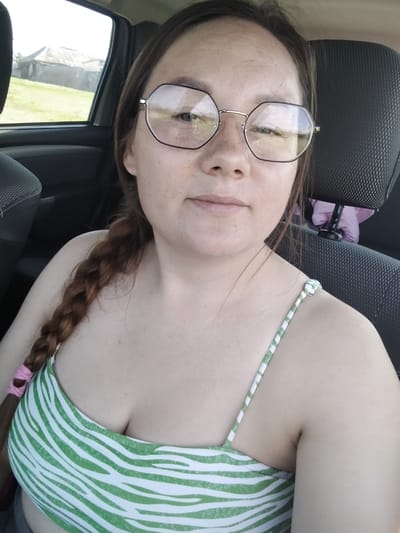 A woman with brown hair in a braid is sitting in the passenger seat of a car. She is wearing glasses with large, hexagonal frames and a green and white striped top. The woman is looking at the camera. The background shows a glimpse of the exterior through the car window, with a building and greenery visible under a bright sky, suggesting it is daytime and likely good weather. The scene appears to be a rural or semi-rural setting in Блохино, Russia. The woman's expression is neutral. There is no visible text in the image.