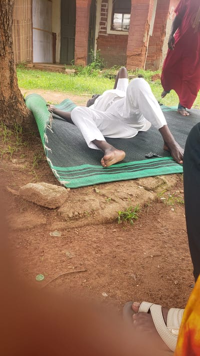 A dark-skinned man wearing white clothing is lying on his back on a green and black mat outdoors. He is positioned on the ground next to a tree trunk. His head is propped up by his hands, and his legs are extended. In the background, a red brick building with arched doorways and a broken window is visible. Green grass and red soil make up the surrounding environment. To the right, a person wearing a red garment and another in black clothing are partially visible. A few small green plants are growing in the dirt.