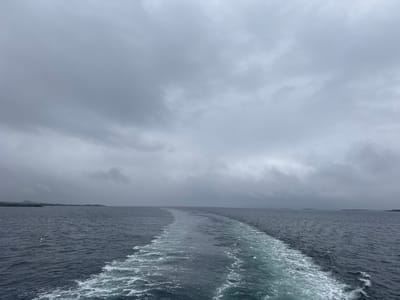 A wide shot captures the open sea under a cloudy sky. The water is a dark, choppy blue. A white wake trails behind the viewer, indicating movement through the water. In the distance, a low, dark landmass is visible, appearing as a hazy silhouette against the horizon. The sky is overcast with dense, grey clouds, suggesting a cool or possibly rainy day. There are no people or specific objects visible in the scene, nor any identifiable landmarks that would suggest the city of Onøya, Norway, without further context. The image depicts a seascape with a focus on the texture of the water and the expansive, muted sky.