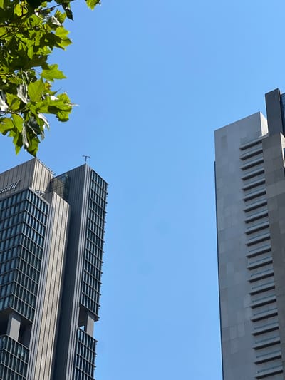 This low-angle shot captures the upper sections of two modern skyscrapers against a clear blue sky. In the foreground, on the left side of the frame, lush green foliage from a tree breaks the composition, its leaves catching the sunlight. The buildings are characterized by their geometric designs and metallic facades. The upper building, positioned at the top of the frame, features a light-colored, segmented facade with a series of dark, rectangular openings, resembling a grand piano keyboard. Below it, another skyscraper dominates the lower right portion of the image. This building has a darker, more angular design, with rows of horizontal windows set within a darker grid-like structure. A distinct architectural detail is the sharp, sawtooth edge along the top of this lower building. The sunlight suggests it is daytime, with clear weather indicated by the absence of clouds. The scene is set in an urban environment, likely a business district, given the presence of such prominent and contemporary architecture. There are no people visible in the image. The overall impression is one of architectural grandeur and a bright, sunny day.