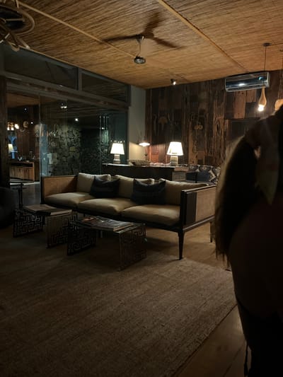 The image depicts a dimly lit interior space with a textured wall on the left composed of horizontal reeds and a wooden accent wall on the right adorned with tribal-like art. An air conditioning unit is mounted high on the wooden wall. A sofa with patterned throw pillows is situated centrally, accompanied by geometric-patterned side tables. A glass partition offers a view of a stone exterior wall and dimly lit interior. The floor is covered with a neutral-toned rug. The scene appears to be a lounge area in a building, potentially a hotel, within Livingstone, Zambia.