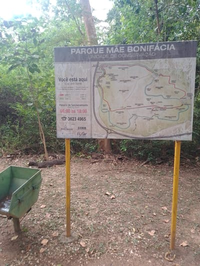 The image depicts a park map at Parque Mãe Bonifácia in Cuiabá, Brazil. The sign is a large, rectangular informational board with a map of the park's trails and points of interest. Text on the sign is in Portuguese, with some English translations. Below the map, there is information about operating hours, a phone number, and logos of organizations such as "Mato Grosso" and "SEMA." The sign is supported by two yellow posts, one of which is partially visible in the foreground. To the left of the sign, there is a green metal trash bin. The ground is covered with dirt, fallen leaves, and small stones. The background consists of lush green trees and foliage, suggesting a natural, outdoor environment. The lighting indicates it is daytime. There are no people visible in the image.