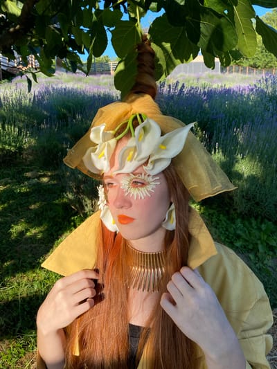 A young woman with long, reddish-brown hair, wearing a yellow collared shirt and a gold necklace, is positioned in the foreground of a lavender field. She is adorned with a yellow veil, white calla lily flowers around her head and ears, and floral eye makeup. Her face is turned to the left, and her hands are resting on her collar. The background consists of a blurred lavender field, green grass, and a wooden fence. The scene is illuminated by the warm light of the sun, casting shadows on the grass.