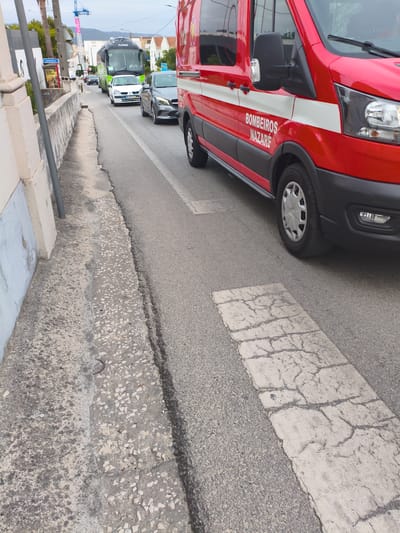 The image shows a street scene in Nazaré, Portugal, on an overcast day. A red fire truck with white stripes and the words "BOMBEIROS NAZARÉ" is prominently featured in the foreground, parked or driving on the right side of the road. The truck has a large side mirror and a part of its front grille and headlight are visible. To the left of the fire truck, a grey car is visible in traffic. Further down the road, a white car and a green bus with "FLIXBUS" written on it are also present in traffic. The road appears to be paved with asphalt and has a white textured stripe marking a pedestrian crossing or speed bump. The left side of the image shows a concrete sidewalk with a textured surface and a wall. The overall environment suggests a town or urban setting. There are no people clearly visible in the image.