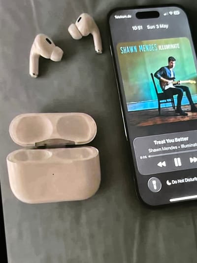 This is a close-up, slightly angled, overhead shot of a smartphone displaying a music player interface and a pair of white wireless earbuds with their charging case. The smartphone screen shows a picture of Shawn Mendes playing a guitar, with the song title "Treat You Better" by Shawn Mendes from the album "Illuminate" visible. The time displayed is 10:51 on Sunday, May 3rd, and "Telekom.de" suggests a German mobile carrier. The earbuds are resting on a grey fabric surface, possibly a bed or couch, along with their open charging case, which reveals two indentations for the earbuds. The scene appears to be indoors, likely in a bedroom, with no indication of the weather or time of day beyond what's on the phone. The overall mood is casual and relaxed, suggesting someone is listening to music. The "Do Not Disturb" icon is also visible on the phone's status bar.