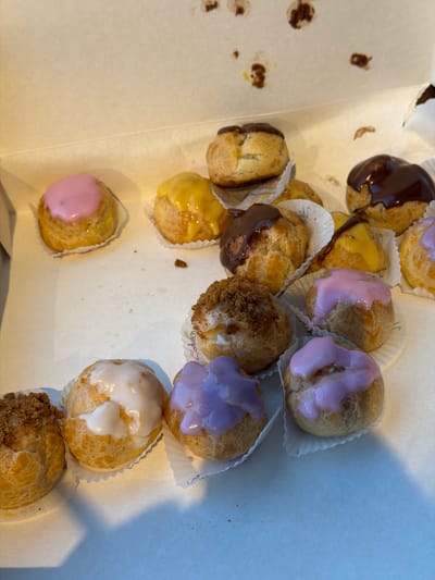 A forensic view of a box of cream puffs, likely in Leudelange, Luxembourg. The box contains several cream puffs with various toppings: chocolate, yellow icing, pink icing, lavender icing, and white icing. Some puffs are in white paper wrappers. Crumbs are scattered in the box. The environment appears to be indoors. No people are visible.