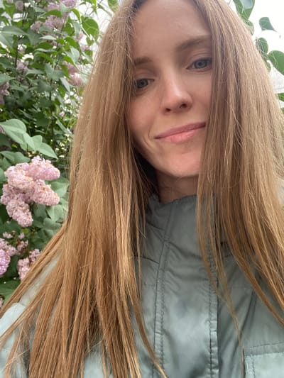 A Caucasian woman with long, blonde hair smiles and looks at the camera. She is wearing a light green jacket. Behind her, there is a bush with pink flowers and green leaves. The image appears to be taken outdoors, possibly in a park or garden.