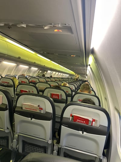 The image shows the interior of an airplane cabin. The seats are arranged in rows, with passengers seated in them. Overhead compartments are visible above the seats. Menus and special deals flyers are tucked into the seatbacks. The airplane appears to be a Norwegian Air Shuttle, based on the branding on the flyers. The image was most likely taken on an aircraft located at the airport in Kastrup, Denmark.