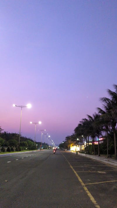 The image captures a street scene in Tuy Hòa, Vietnam, at twilight. The sky is a gradient of purple hues. Palm trees line the left side of the frame, bordering the road. Streetlights illuminate the road, stretching into the distance. A motorcycle with red taillights is visible further down the road. The road itself is dark asphalt with painted yellow lines. Buildings and greenery are present along the roadside.