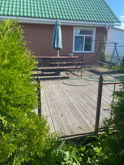 This is a high-angle, full shot of a wooden deck in front of a brick house in Bol'shoe Zaborod'e, Russia. The deck is empty except for a green garden hose coiled on the ground. To the left, a closed teal umbrella stands on a patio table with two benches, a small potted plant, and a cup. Behind the table and benches, a green metal swing set is visible. A large green evergreen tree is positioned in the foreground on the left, and another smaller one is on the right, partially obscuring the view of the deck and the house. The house has a green metal roof, a white gutter, and a single window with a white frame. The sky is blue with some clouds.