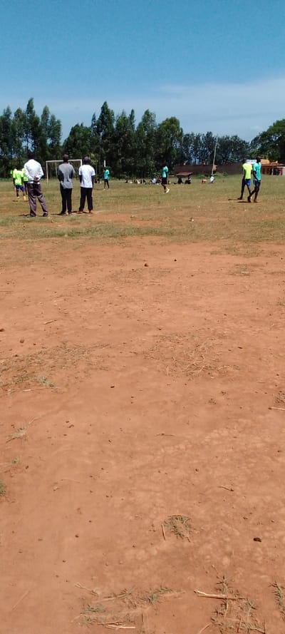 The image shows a dirt field with a few scattered tufts of green grass and some small, brown, dry twigs. In the background, there is a line of trees that have lost their leaves. In the middle distance, there are a few figures running on the field. The figures are wearing a mix of light blue and green shirts. There is a small goalpost to the left of the frame. The image appears to have been taken on a sunny day in Kipkarren, Kenya, with clear skies. The location is approximately 0.613 latitude, 34.975 longitude.