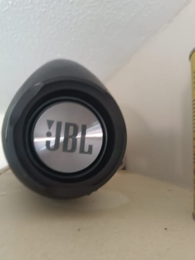 A close-up, low-angle shot focuses on a black JBL portable speaker positioned on a light-colored, textured surface, possibly a shelf. The speaker's circular grille is prominently displayed, featuring the silver JBL logo in bold, sans-serif font. The background consists of a white, textured wall or ceiling meeting a neutral-colored wall. To the right of the speaker, a portion of a yellow object, possibly a can or marker, is visible. The scene appears to be an indoor setting. There is no indication of the specific location within Huntingdonshire, United Kingdom.
