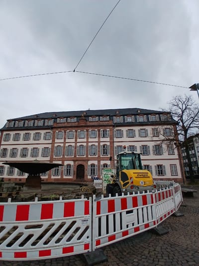 The image shows a construction site in front of a large, ornate building in Darmstadt, Germany. The building is several stories high, with a red and white facade and numerous windows. A bare tree is visible to the left of the building. A yellow construction vehicle, possibly a small excavator, is parked on the site, near a barrier made of red and white striped panels. The ground is paved with cobblestones. Overhead, power lines cross the gray sky.