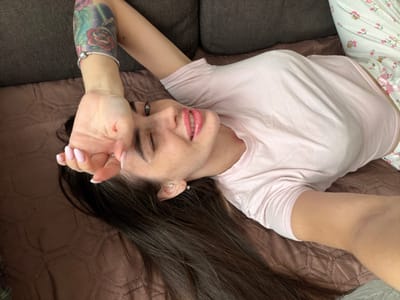 The scene depicts a female individual lying in an indoor residential setting, ostensibly within the city of Novosaratovka, Russia. The individual, an adult with long dark brown hair, is positioned with her head towards the upper left of the frame, looking directly towards the camera. Her face features freckles, and she wears light pink lipstick. Her left eye is partially closed, while her right eye is open. A silver stud earring is visible in her left ear. Her left arm is raised, with her hand partially covering her forehead. The nails on her visible left hand display a pink and white French manicure. A silver charm bracelet is present on her left wrist. A colorful tattoo featuring red roses and green foliage is visible on her left upper arm. She is attired in a light pink short-sleeved t-shirt and white pajama bottoms with small pink and green floral patterns. She is lying on a surface covered by a dark brown, quilted blanket or throw with a diamond pattern. A dark gray sofa cushion is visible behind her head.