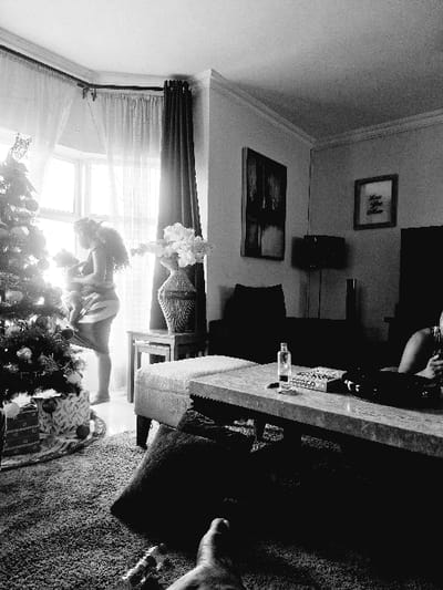 A black and white indoor shot reveals a room with a partial view of a Christmas tree in the foreground, likely positioned near a window with sheer curtains. A woman is seen bending over, potentially arranging gifts or decorations. The room contains a sofa and a coffee table on which a bottle is placed. Pictures adorn the walls. The ceiling has decorative moldings. Part of a person's foot is visible near the carpet.
