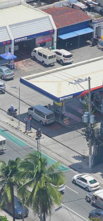 The image depicts an aerial view of a street scene in Makati, Philippines, dominated by a Petron gas station. Several vehicles are present, including white vans, a white SUV, a silver sedan, a blue sedan, a white taxi, and a jeepney. A person on a motorcycle is seen near the gas pumps. The gas station features a prominent "PETRON" sign and several fuel pumps. Adjacent to the gas station is a "SERVICE" building, possibly an auto repair shop, with a blue canopy and a sign advertising various services. Palm trees line the street, and a bicycle lane is visible. The overall environment appears to be an urban setting with a mix of commercial and residential buildings in the background. The sky is bright, suggesting a daytime scene.