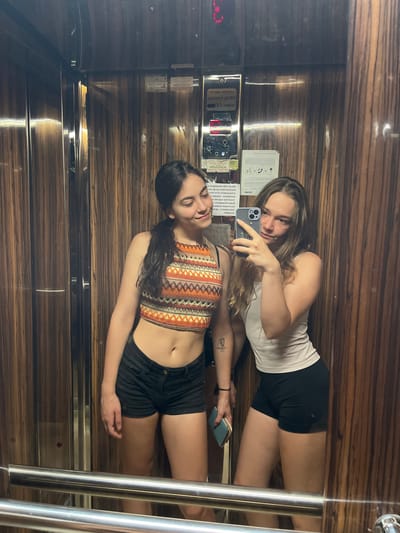 The image shows two young women taking a selfie in an elevator. The elevator's walls are paneled with a dark wood grain pattern, and there are metal handrails in the foreground. The woman on the left has dark hair pulled back in a ponytail and is wearing a patterned crop top and black shorts. She is looking towards the camera with a slight smile. The woman on the right has blonde hair and is wearing a white tank top and black shorts. She is holding a smartphone and appears to be taking the selfie. The background shows elevator control panels and signage, which is consistent with an elevator in a building. The presence of a sign with Cyrillic script, potentially indicating a location in Bulgaria, is noted. Given the information provided, this elevator could be located in Lozenets, Bulgaria, a popular resort town.