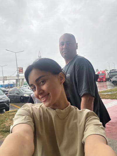 In a photograph taken in the city of Argveta, Georgia, a young woman with dark hair smiles at the camera while holding it to take a selfie. Behind her, a bald man in a dark t-shirt stands, looking towards the camera. In the background, a street scene unfolds with various vehicles, including cars and large trucks. The sky is overcast, suggesting cloudy weather. The setting appears to be an outdoor public area, possibly a parking lot or roadside, with signs and streetlights visible. The ground is wet, indicating recent rain.