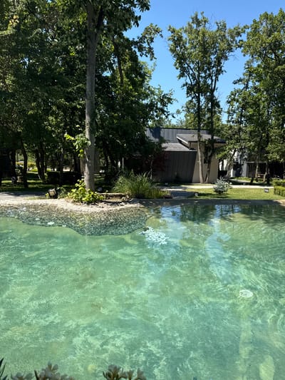 The image shows a clear turquoise swimming pool with pebbles visible on the bottom. In the background, there is a modern house with a dark, angular roof, surrounded by lush green trees. The sky is a clear blue. The pool is situated in a garden-like setting, suggesting it is part of a private residence or a resort. The overall atmosphere is serene and peaceful, with the water reflecting the surrounding greenery and the blue sky. There are no people visible in the image. The location of Atiya, Bulgaria is not explicitly indicated in the image itself, but the description assumes it is part of a known location.