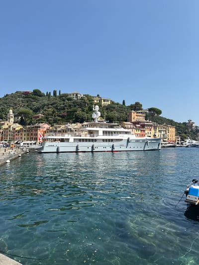 This image depicts a harbor in Portofino, Italy, on a sunny day. A large, light blue luxury yacht is docked in the foreground, its name "ELISABET" visible on its hull. The yacht has multiple decks, numerous windows, and is adorned with safety buoys. Behind the yacht, the colorful buildings of Portofino line the waterfront, nestled against a verdant, hilly landscape dotted with trees and houses. A prominent bell tower of a church rises above the town. The water in the harbor is clear and blue, with sunlight dappling its surface, revealing the seabed below in some areas. Several smaller boats are moored in the background. A few people are visible on the docks, and a small utility boat with a blue container is tied off to the right side of the frame. The sky is a clear, bright blue.